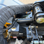 Close-up of heavy-duty hydraulic/mast assembly with hoses, bolts, and a McConnel label, showing mechanical linkages and protective padding on a yellow-and-black machine.