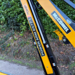 Close-up of a yellow hydraulic excavator arm with black and gray decals, leaning against a leafy hedge.