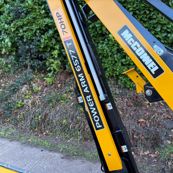 Close-up of a yellow hydraulic excavator arm with black and gray decals, leaning against a leafy hedge.