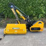 Yellow McConnel construction attachment with hydraulic arms resting on the ground, hoses trailing forward, hedge in the background.