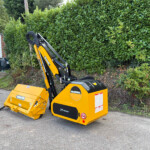 Yellow mini excavator with its front bucket raised, parked beside a tall green hedge near a storefront and a red car in the background.