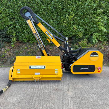 Yellow McConnel construction machine with a hydraulic arm and black hoses, parked on pavement in front of a green hedge.