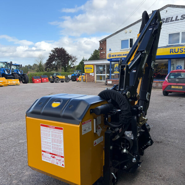 Close-up of a yellow McConnel agricultural attachment in the foreground at a farm equipment dealer, with blue tractors and a Russell's sign visible in the background on the building.