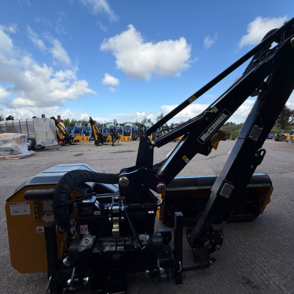Close-up of a black hydraulic arm labeled 'McConnel' on an orange construction machine in a yard, with pallets and other equipment in the background under a blue sky with clouds.