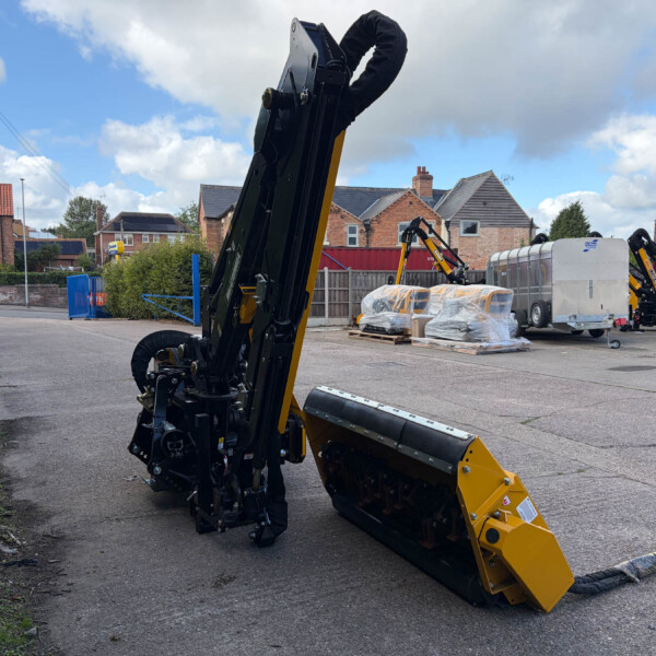 Large yellow and black tracked construction machine in a yard, with wrapped pallets and brick houses in the background