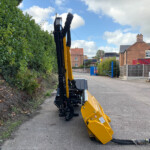 Yellow construction excavator extending its arm along a street beside a tall hedge, with houses in the background.