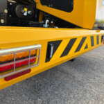 Back of a bright yellow utility truck with black hazard stripes and red/amber tail lights on a paved street.