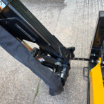 Close-up of a black metal forklift attachment with a hydraulic pin, yellow frame, and safety label on pavement.