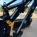 Close-up of a black hydraulic machine arm with McConnel branding and yellow components, in a tractor yard background of blue skies and equipment.