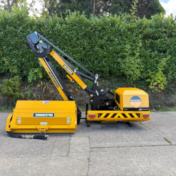 Yellow McConnell construction machine with hydraulic arms parked on a paved lot in front of a dense green hedge.