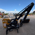 Large black hydraulic crane arm in foreground at a construction yard, with pallets, a yellow generator, and blue sky.