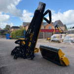 Construction machinery with a long black arm in a yard; pallets wrapped in plastic and brick houses in the background