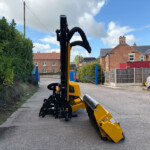 Yellow construction machine with an extended articulated arm on a residential street, hedge on the left and brick houses in the background.