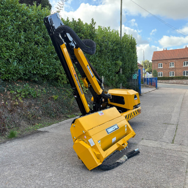 Yellow compact construction machine with a folded black arm resting on a paved road beside a tall green hedge and a brick building in the background.