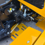 Close-up of yellow construction machine frame exposing hydraulic pump, hoses, and fittings in a black engine area.