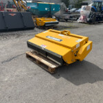 Yellow McConnel agricultural mower on a wooden pallet in an outdoor equipment yard, with various machines and a blue New Holland tractor in the background.