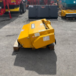Yellow industrial machinery sits on a wooden pallet in a gravel lot, with red and teal equipment in the background.