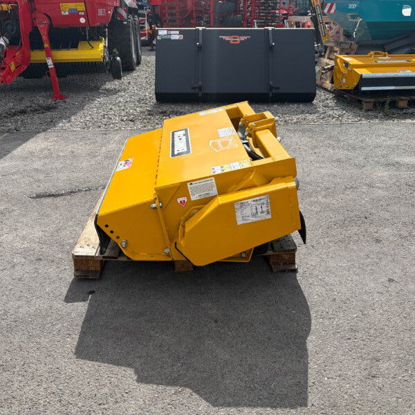 Yellow industrial machinery sits on a wooden pallet in a gravel lot, with red and teal equipment in the background.