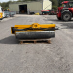 Yellow road roller sits on a wooden pallet in an outdoor industrial yard with a beige metal warehouse, a white van labeled Russell’s Agriculture, and a red Manitou telehandler in the background.