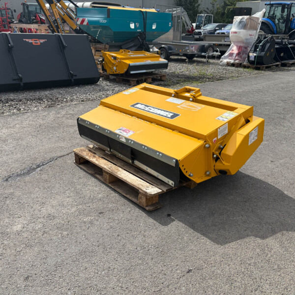 Yellow McConnel agricultural mower on a wooden pallet in an outdoor equipment yard, with various machines and a blue New Holland tractor in the background.