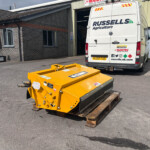 Rear view of a white van with Russell Agriculture logo parked outside a workshop, with yellow machinery in the foreground on pallets.