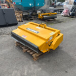 Yellow McConnel industrial attachment on a wooden pallet in a gravel lot, with other construction machinery and a teal excavator in the background.