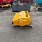 Yellow metal industrial attachment on a wooden pallet in a gravel lot, with large red and teal machines in the background.
