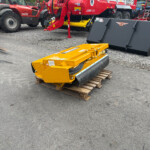 Yellow industrial roller attachment placed on a wooden pallet in a gravel lot, with red heavy machinery in the background.