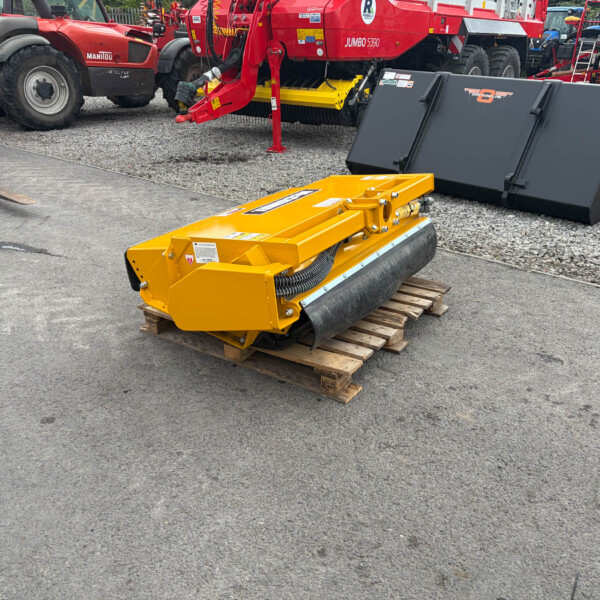 Yellow industrial roller attachment placed on a wooden pallet in a gravel lot, with red heavy machinery in the background.