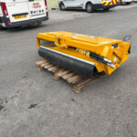 Yellow road roller/compactor resting on wooden pallets in a parking lot with white vans in the background.