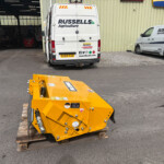 Yellow industrial machine on wooden pallets in a parking lot with white vans labeled Russell's Agriculture in the background.