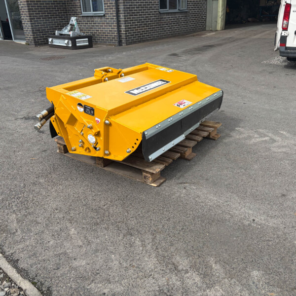 Yellow industrial machine attachment on a wooden pallet in a paved outdoor lot, with a white van and brick building in the background.