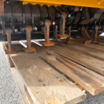 Rusty undercarriage of agricultural equipment with rusted blades and bolts, resting on a wooden pallet beneath a yellow frame.