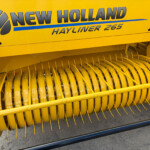 Close-up of a bright yellow New Holland Hayliner 265 harvester header, showing curved metal tines and the pickup area at the bottom.