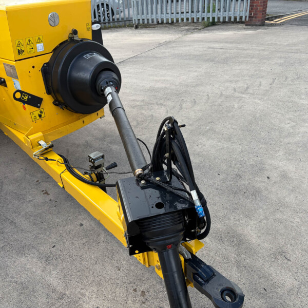 Close-up of a yellow industrial machine with a large black hydraulic component and cables, on a paved surface near a fence.