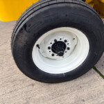 Close-up of a heavy-duty black tire with a white rim, mounted on yellow industrial equipment behind it on concrete.]","Wheel and tire: black treaded tire with white rim, part of yellow machinery visible in the background.