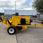 Bright yellow Hayliner 265 hay baler machine parked in a fenced outdoor lot with an industrial corrugated metal building in the background.