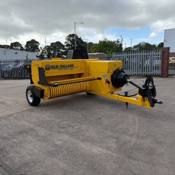 Yellow New Holland Hayliner 265 baler parked in a concrete yard with a metal fence and industrial buildings in the background.