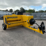Yellow New Holland Hayliner 265 baler parked in an industrial lot with a metal fence behind it.