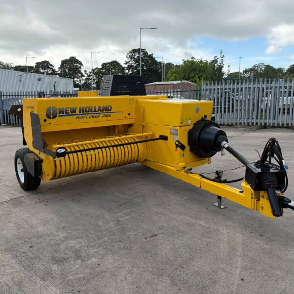Yellow New Holland Hayliner 265 baler parked in an industrial lot with a metal fence behind it.