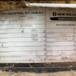Close-up of a dirt-covered machinery data plate showing CNH Industrial Belgium and New Holland Agriculture branding and model information.