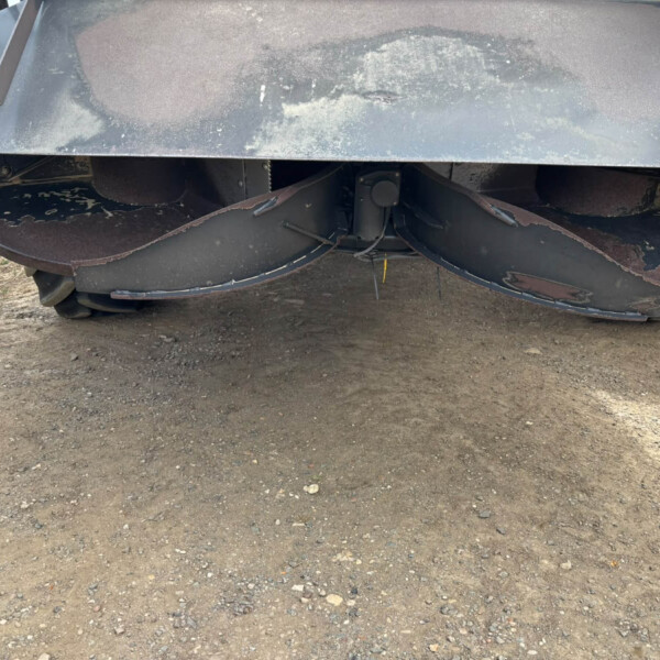 Underside of a large, rusted agricultural mower with circular blades, dirt and gravel ground beneath it.
