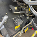 Close-up of heavy construction machinery showing a black metal frame, hydraulic hoses, bolts, and wires with yellow caution labels on the lower panel