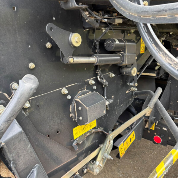 Close-up of heavy construction machinery showing a black metal frame, hydraulic hoses, bolts, and wires with yellow caution labels on the lower panel