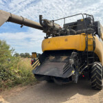 Large yellow construction machine with a long discharge chute extending to the left, on a dirt road with bushes and a blue-sky backdrop.