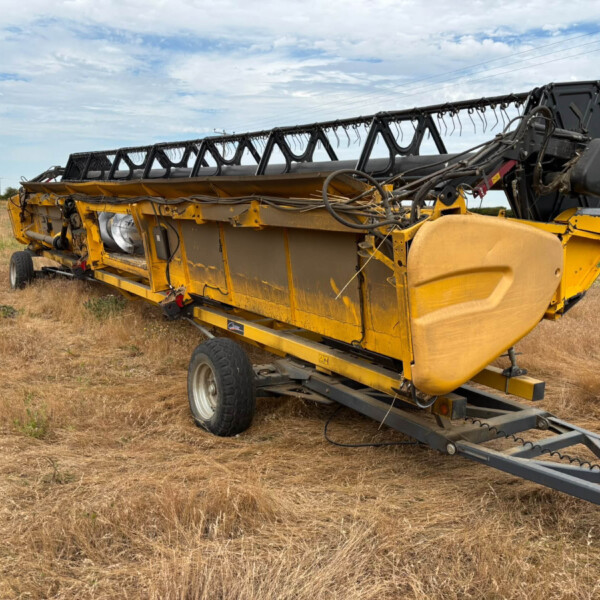 Large yellow harvesting header lying on its side in a dry field under a partly cloudy blue sky.