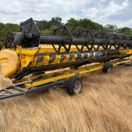 Yellow and black combine harvester header on a trailer in a dry grassy field with trees in the background.