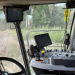 Tractor cab interior with dual screens, steering wheel, and control console; field greenery visible outside the window.