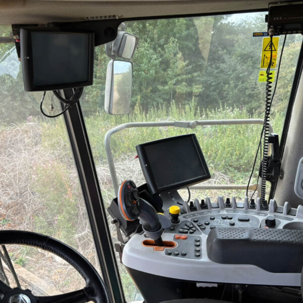 Tractor cab interior with dual screens, steering wheel, and control console; field greenery visible outside the window.