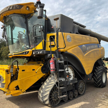 Yellow New Holland combine harvester with tracked wheels and front header on a dirt road, under a cloudy sky